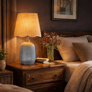May include: A bedside lamp with a textured blue base and a beige lampshade. The lamp sits on a wooden nightstand with a drawer. A vase of flowers and a framed picture are in the background. The lamp is a decorative lighting fixture.