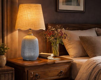 May include: A bedside lamp with a textured blue base and a beige lampshade. The lamp sits on a wooden nightstand with a drawer. A vase of flowers and a framed picture are in the background. The lamp is a decorative lighting fixture.
