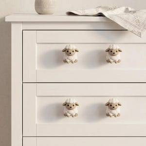 May include: White dresser with six sheep-shaped drawer pulls. The sheep are white with tan faces, ears, and feet. A vase with dried plants and a folded cloth are on top of the dresser.
