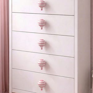May include: A white chest of drawers with five drawers, each featuring a hot air balloon-shaped knob. The knobs are white with pink stripes. The chest of drawers is in a room with pink accents.