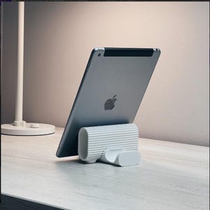 May include: A silver tablet with the Apple logo is supported by a white, ribbed tablet stand. The stand has a curved base and a cylindrical body. The background is a light, neutral colour.