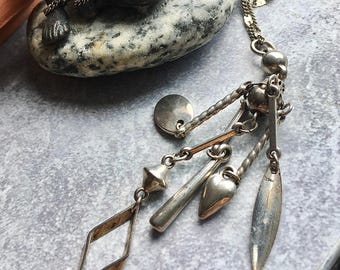 May include: A silver-toned necklace featuring a delicate chain and a cluster of dangling charms. The charms include a flat circular disc, a twisted metal bar, a smooth heart shape, a slender leaf-like pendant, and an open diamond-shaped frame.