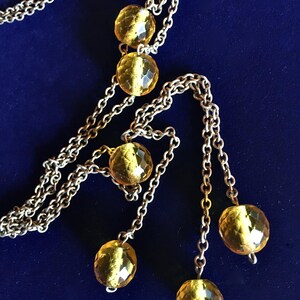 May include: A silver-toned necklace with multiple strands and dangling faceted amber-colored beads. The necklace is displayed against a dark blue background, highlighting the jewelry's design and color.