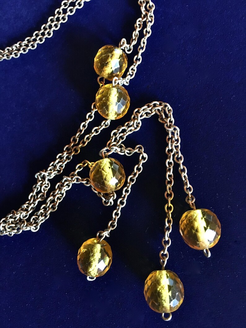 May include: A silver-toned necklace with multiple strands and dangling faceted amber-colored beads. The necklace is displayed against a dark blue background, highlighting the jewelry's design and color.