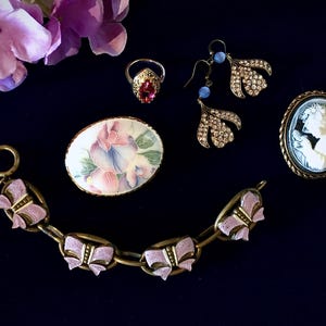 Può includere: Un assortimento di gioielli vintage, tra cui un anello con pietra rossa, orecchini con perline blu, una spilla cammeo e un braccialetto con dettagli a fiocco rosa. I gioielli sono disposti su uno sfondo blu scuro.