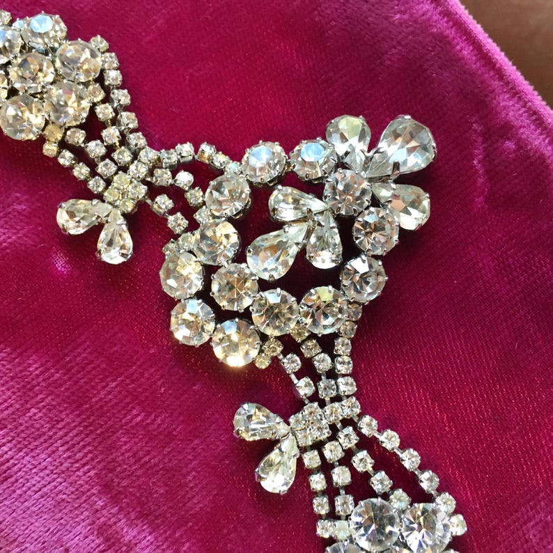 May include: A silver-toned necklace featuring clear, faceted rhinestones arranged in floral and teardrop patterns. The design includes multiple circular and pear-shaped stones connected by delicate metal links, creating a shimmering, intricate jewelry piece.
