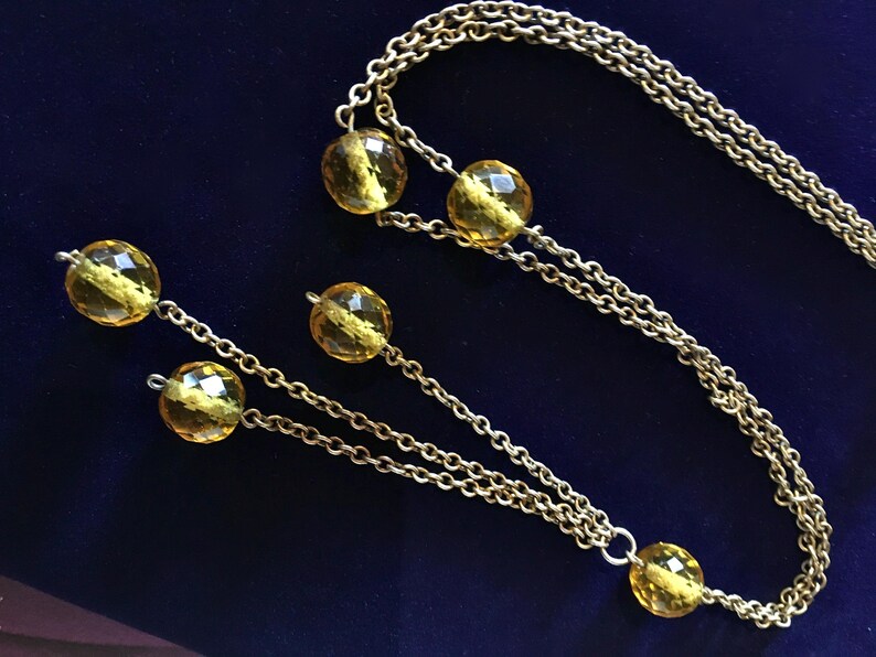 May include: A gold-toned necklace with faceted yellow beads. The necklace features a long chain with multiple strands and decorative beads. The beads are evenly spaced along the chain, creating a classic and elegant look.