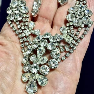 May include: A silver-toned metal necklace and earring set featuring clear, faceted rhinestones arranged in floral and teardrop patterns. The jewelry pieces are constructed with multiple rows of small, round stones connecting larger, brilliant-cut gems.