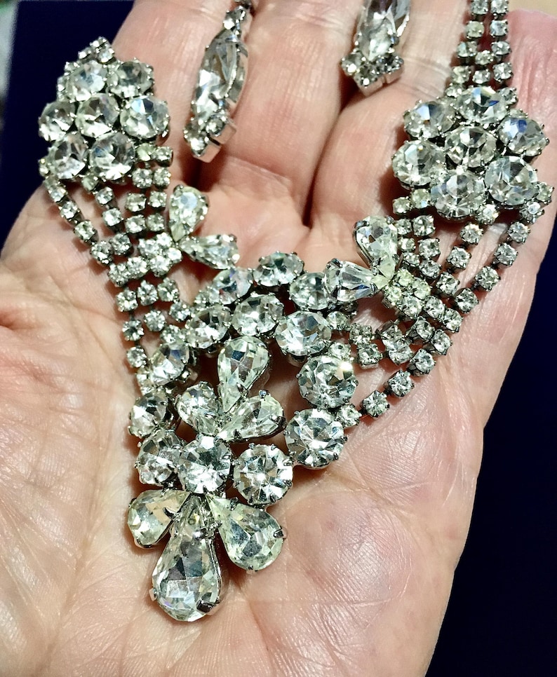 May include: A silver-toned metal necklace and earring set featuring clear, faceted rhinestones arranged in floral and teardrop patterns. The jewelry pieces are constructed with multiple rows of small, round stones connecting larger, brilliant-cut gems.