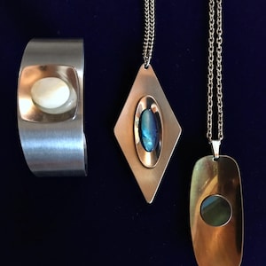 May include: Three silver and gold tone necklaces with oval shaped stones. The stones are iridescent and have a blue and green colour. The necklaces are on a dark blue background.