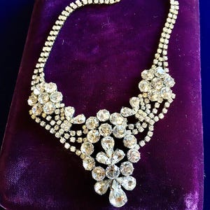May include: A silver-tone metal necklace featuring multiple rows of clear, faceted rhinestones. The design includes two floral-shaped clusters on the sides and a central, elongated pendant element composed of various round and pear-shaped stones.