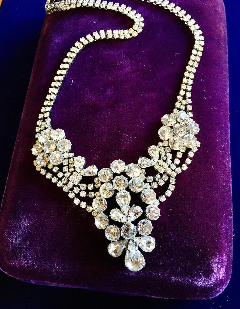 May include: A silver-tone metal necklace featuring multiple rows of clear, faceted rhinestones. The design includes two floral-shaped clusters on the sides and a central, elongated pendant element composed of various round and pear-shaped stones.