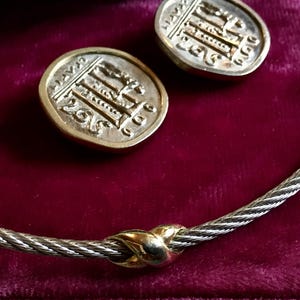 Puede incluir: Un collar plateado con un diseño de cable trenzado y detalles dorados, junto con dos pendientes redondos a juego. Los pendientes y el collar se exhiben sobre una superficie de terciopelo rojo oscuro. Los pendientes tienen un borde dorado y un diseño detallado.