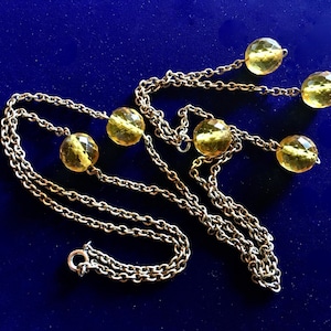 May include: A vintage necklace with a silver chain and six faceted, yellow-gold beads. The beads are evenly spaced along the chain, with three beads on each side. The necklace is displayed on a dark blue background.