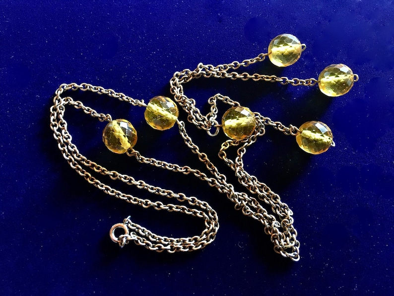 May include: A vintage necklace with a silver chain and six faceted, yellow-gold beads. The beads are evenly spaced along the chain, with three beads on each side. The necklace is displayed on a dark blue background.