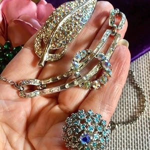 May include: A silver-tone metal leaf brooch covered in iridescent rhinestones, a curved necklace segment featuring baguette and round iridescent rhinestones, and a round cluster ring set with small blue and iridescent rhinestones.