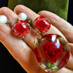 Könnte beinhalten: Ein Set Vintage-Schmuck mit roten Rosen, die in klarem Glas eingeschlossen sind. Die Ohrringe sind herzförmig und haben einen Perlenakzent. Der Anhänger ist tropfenförmig.