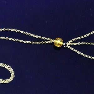 May include: A delicate gold-colored necklace with multiple strands and yellow gemstone accents. The necklace features a chain design with several strands extending from a central point, each ending with a round, yellow gemstone. The necklace is displayed on a dark blue surface.