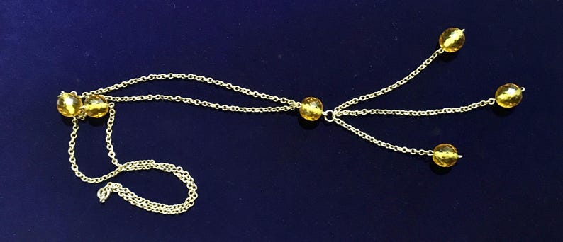 May include: A delicate gold-colored necklace with multiple strands and yellow gemstone accents. The necklace features a chain design with several strands extending from a central point, each ending with a round, yellow gemstone. The necklace is displayed on a dark blue surface.