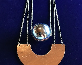May include: A silver-toned necklace featuring a large, reflective sphere pendant suspended from a delicate chain. Below the sphere is a copper-colored, crescent-shaped pendant. The necklace is displayed against a dark blue background.