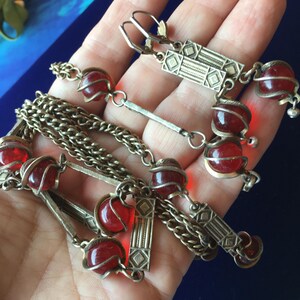 Könnte beinhalten: Ein Halsketten- und Ohrringset mit antikfarbenen Metallketten und rechteckigen geometrischen Gliedern. Der Schmuck enthält durchscheinende rote, kugelförmige Perlen, die in zarte Drahtkäfige gefasst sind und ein einheitliches Design bilden.