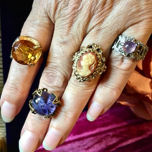 Könnte beinhalten: Vier Ringe an den Fingern: einer mit einem großen facettierten bernsteinfarbenen Stein, einer mit einem runden violetten Stein in dekorativer Fassung, einer mit einem geschnitzten Kamee-Porträt und einer mit einem kleinen runden lavendelfarbenen Stein.