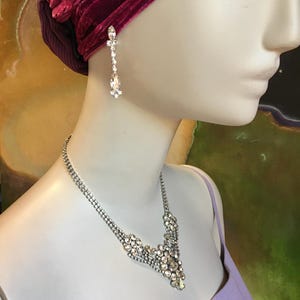 May include: A silver-toned necklace and matching drop earring set featuring clear, faceted crystals. The necklace has a central cluster of stones, while the earring consists of a small crystal stud connected to a larger, teardrop-shaped crystal.