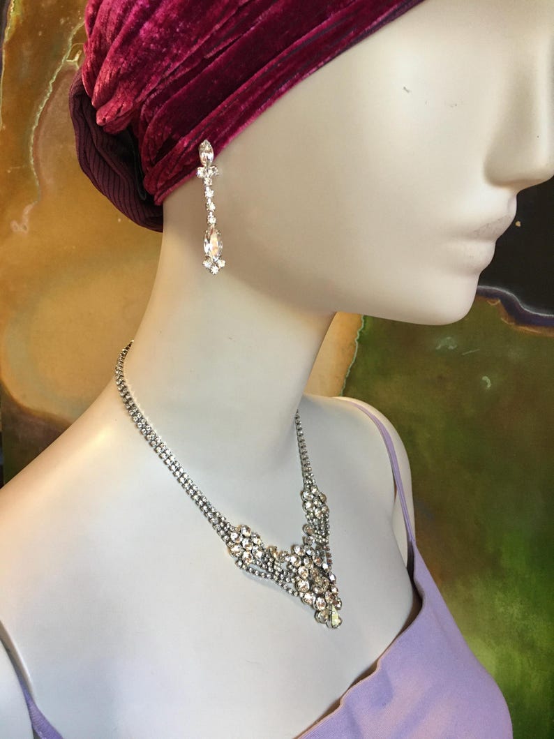 May include: A silver-toned necklace and matching drop earring set featuring clear, faceted crystals. The necklace has a central cluster of stones, while the earring consists of a small crystal stud connected to a larger, teardrop-shaped crystal.