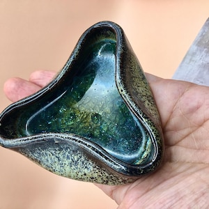 Può includere: Posacenere in ceramica fatto a mano con una forma triangolare unica. L'interno presenta una miscela di tonalità verdi e blu, mentre l'esterno ha una finitura strutturata e macchiettata. Il bordo del posacenere è scuro.