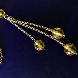 May include: A gold-toned necklace with multiple strands and faceted yellow beads. The necklace has a delicate chain design and a dark blue background.
