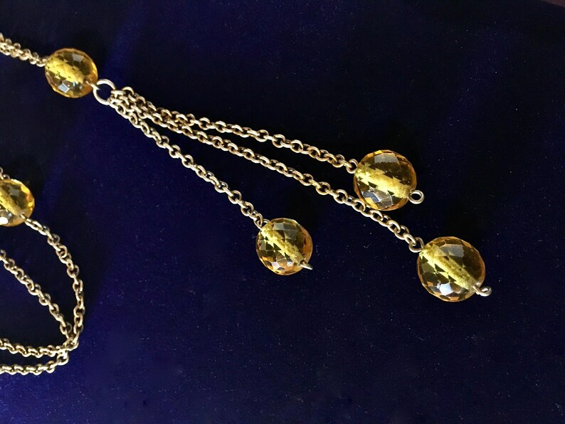 May include: A gold-toned necklace with multiple strands and faceted yellow beads. The necklace has a delicate chain design and a dark blue background.