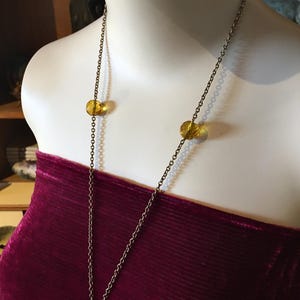May include: A long, antique gold-tone necklace with three amber-colored, round beads. The necklace is displayed on a mannequin wearing a burgundy velvet top. The chain is delicate and hangs down the front.
