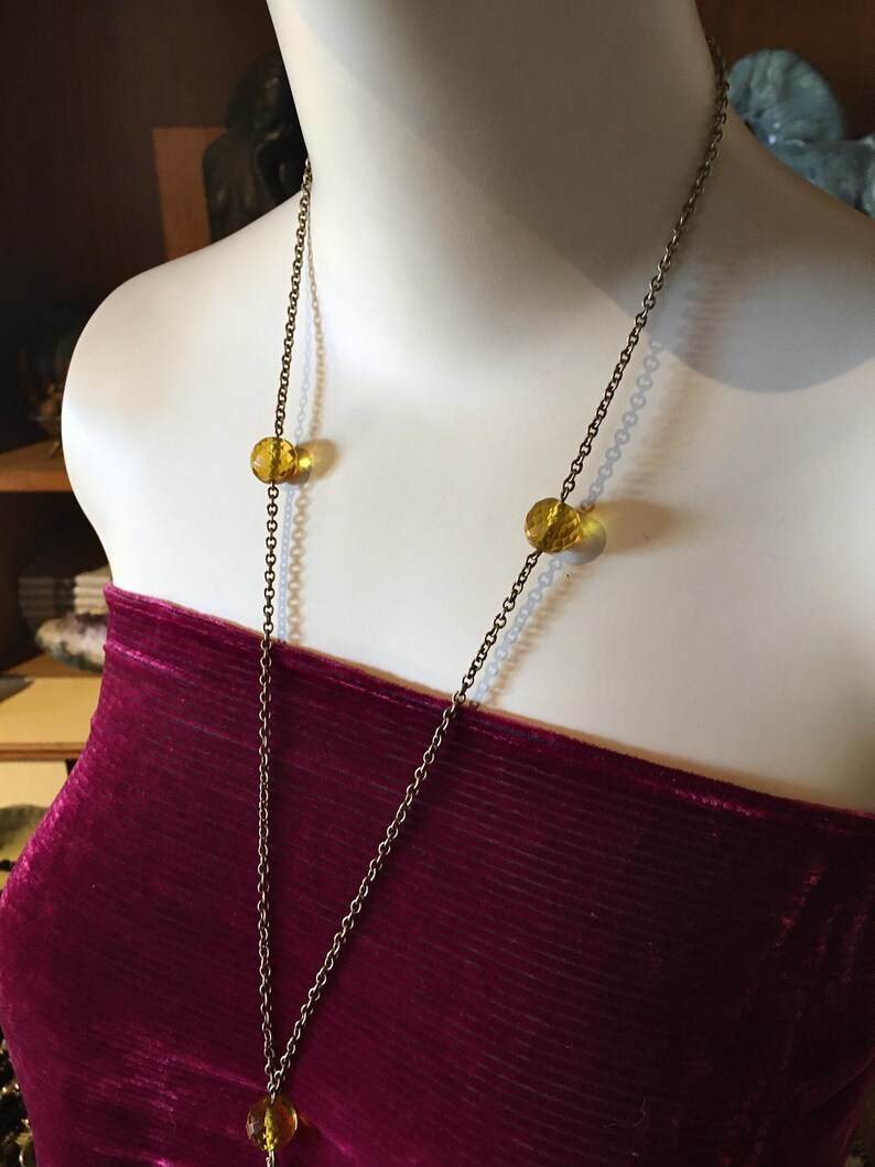 May include: A long, antique gold-tone necklace with three amber-colored, round beads. The necklace is displayed on a mannequin wearing a burgundy velvet top. The chain is delicate and hangs down the front.