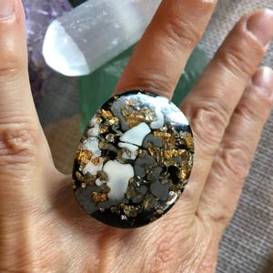 Könnte beinhalten: Ein ovaler Ring mit schwarzer Basis, der ein Mosaik aus weißen, grauen und goldfarbenen Akzenten aufweist. Das Design des Rings ist auffällig, wobei die Goldelemente einen Hauch von Luxus verleihen. Der Ring ist ein Statement-Schmuckstück.