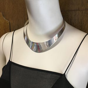 Może przedstawiać: Polerowany naszyjnik typu choker z metalu w kolorze srebrnym z szerokim, zakrzywionym panelem przednim w kształcie półksiężyca, który zwęża się ku tyłowi. Gładka, odblaskowa powierzchnia kołnierza ma wysoki połysk i spoczywa na białym manekinie.