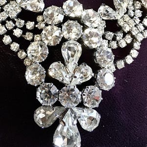 May include: A silver-toned necklace featuring multiple strands of clear, faceted rhinestones. The central pendant design consists of a cluster of round and pear-shaped stones arranged in a symmetrical pattern, connected to layered rhinestone chains.