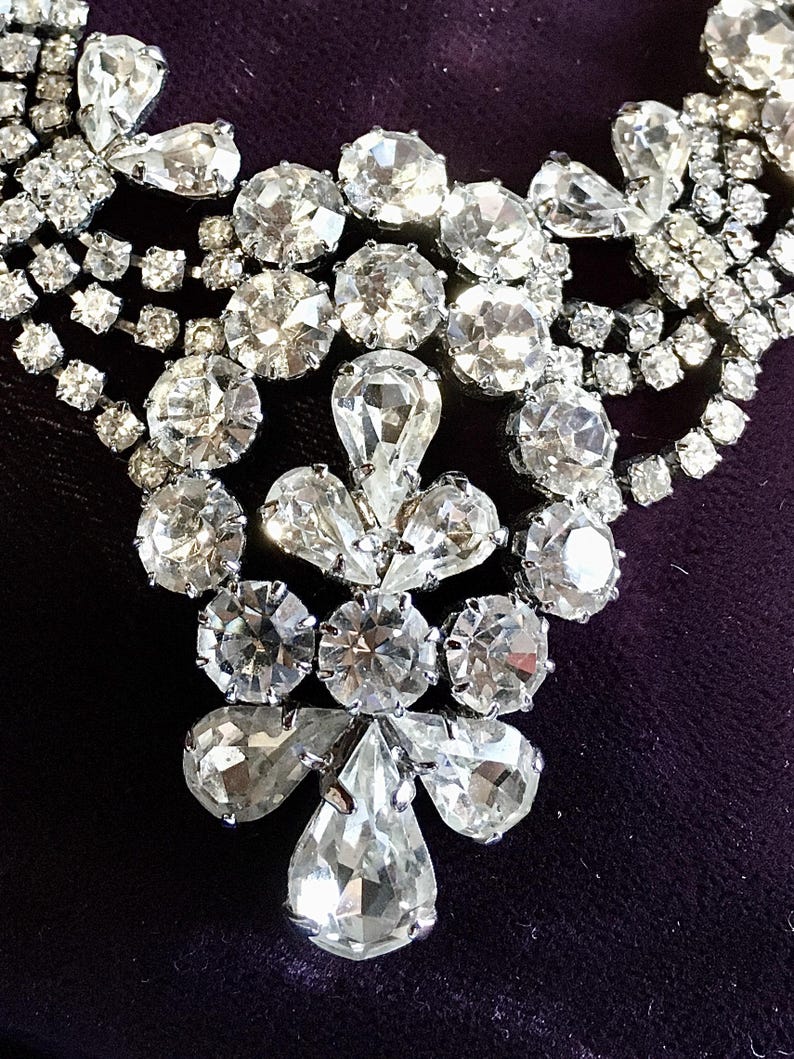 May include: A silver-toned necklace featuring multiple strands of clear, faceted rhinestones. The central pendant design consists of a cluster of round and pear-shaped stones arranged in a symmetrical pattern, connected to layered rhinestone chains.