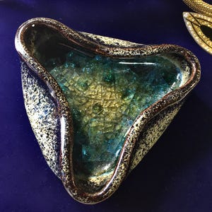 Può includere: Un posacenere triangolare in ceramica con esterno maculato nei toni del beige, blu e marrone. L'interno presenta un mosaico di pezzi di vetro blu e verde. Il bordo è di colore marrone scuro.