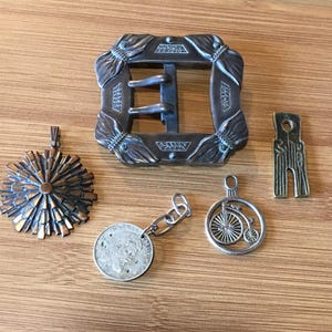 Pode incluir: Uma coleção de acessórios de metal incluindo uma fivela de cinto retangular grande com detalhes ornamentados, um pingente circular em forma de sol, uma moeda presa a uma pequena corrente de metal, um pingente de bicicleta e uma etiqueta metálica.