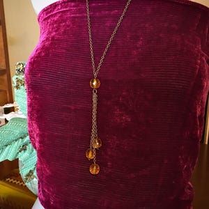 May include: A gold-toned necklace with amber-colored beads. The necklace has a long chain with a pendant of three dangling beads. The necklace is displayed on a burgundy velvet top.