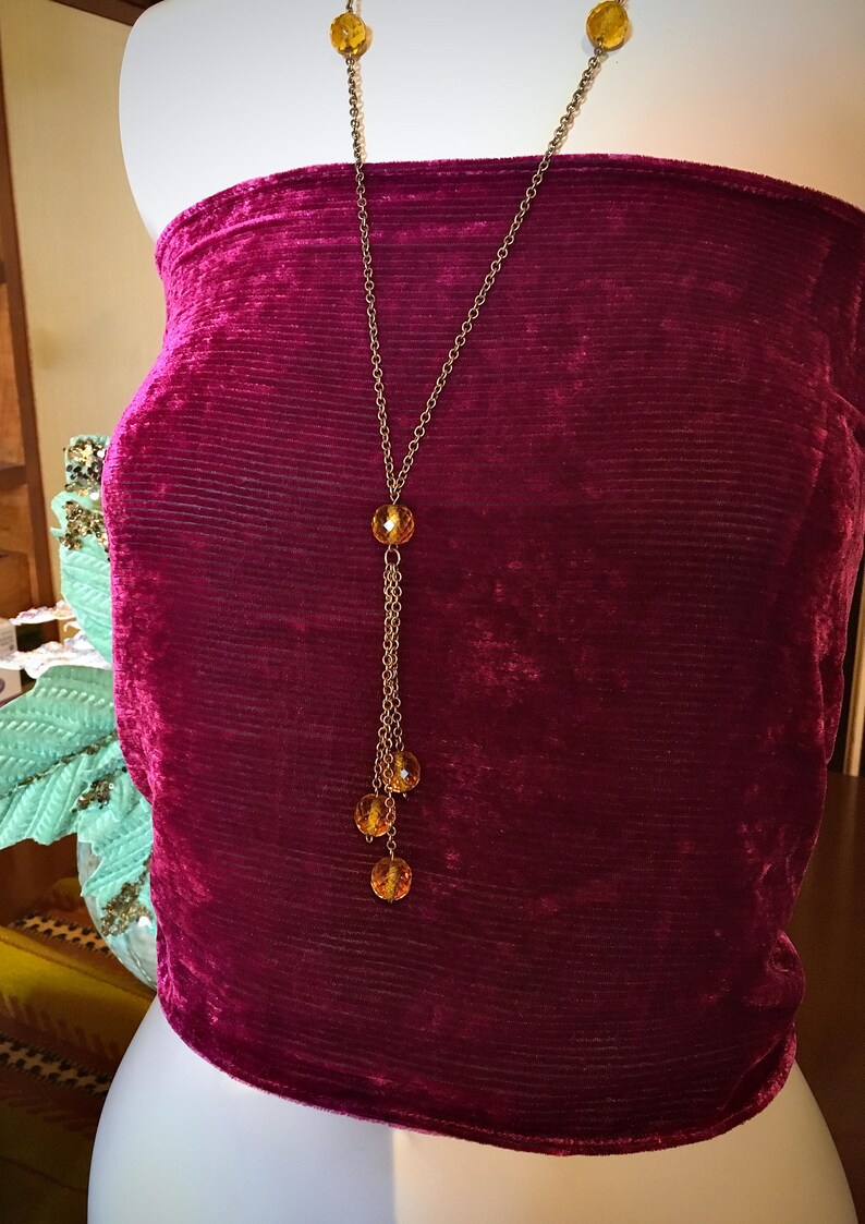 May include: A gold-toned necklace with amber-colored beads. The necklace has a long chain with a pendant of three dangling beads. The necklace is displayed on a burgundy velvet top.