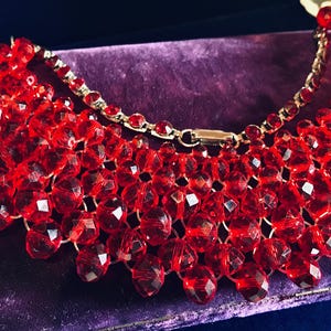Peut inclure: Un collier de perles rouge vif avec un design superposé. Le collier présente de nombreuses perles à facettes, créant un effet scintillant. Le collier a une chaîne et un fermoir dorés, reposant sur une surface en velours violet.