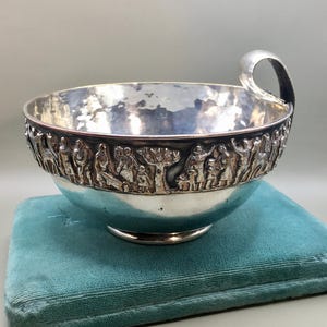 Puede incluir: Un cuenco de plata con interior pulido y textura martillada. El exterior presenta un friso en relieve continuo que representa figuras, árboles y animales. El cuenco incluye un asa curva única fijada en el borde superior.