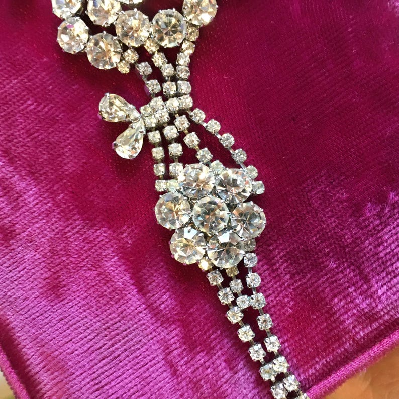 May include: A silver-toned metal bracelet featuring clear, faceted rhinestones arranged in a multi-strand design. The piece includes a central floral cluster of round stones and a smaller, teardrop-shaped accent, all set in a delicate, articulated chain.