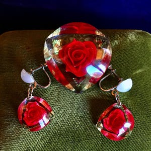 Può includere: Un set vintage di spilla e orecchini a forma di rosa rossa. La spilla e gli orecchini sono realizzati in plastica trasparente con una rosa rossa incastonata all'interno. Gli orecchini sono di tipo clip.