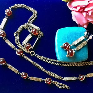 Könnte beinhalten: Ein Set aus einer goldfarbenen Metallkette und passenden Ohrhängern mit rechteckigen, gravierten Gliedern und runden, durchscheinenden dunkelroten Glasperlen, die mit dünnem Draht umwickelt sind. Der Schmuck ist mit einer türkisfarbenen Schachtel arrangiert.