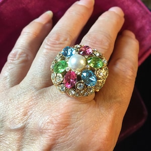 Puede incluir: Un anillo dorado con una perla blanca central rodeada de piedras preciosas de colores rosa, verde y azul. El anillo está adornado con pequeños cristales transparentes, creando un efecto brillante. Un anillo llamativo.