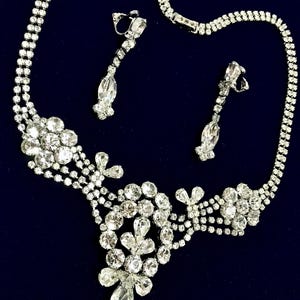 May include: A silver-toned necklace and matching clip-on earring set featuring clear, faceted rhinestones. The necklace has a multi-strand chain design with floral-shaped clusters and teardrop-shaped stones. The earrings are long, linear drops.