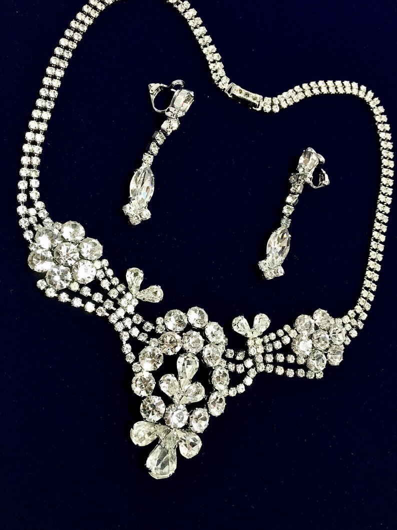 May include: A silver-toned necklace and matching clip-on earring set featuring clear, faceted rhinestones. The necklace has a multi-strand chain design with floral-shaped clusters and teardrop-shaped stones. The earrings are long, linear drops.