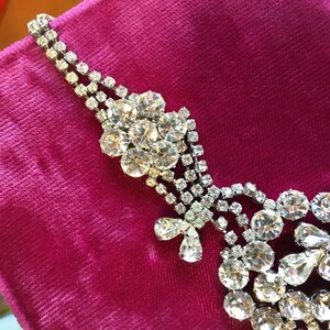 May include: A silver-toned necklace featuring multiple strands of clear, round-cut rhinestones. The design includes a floral-shaped cluster of stones and a dangling element composed of pear-shaped and round-cut rhinestones set in metal.